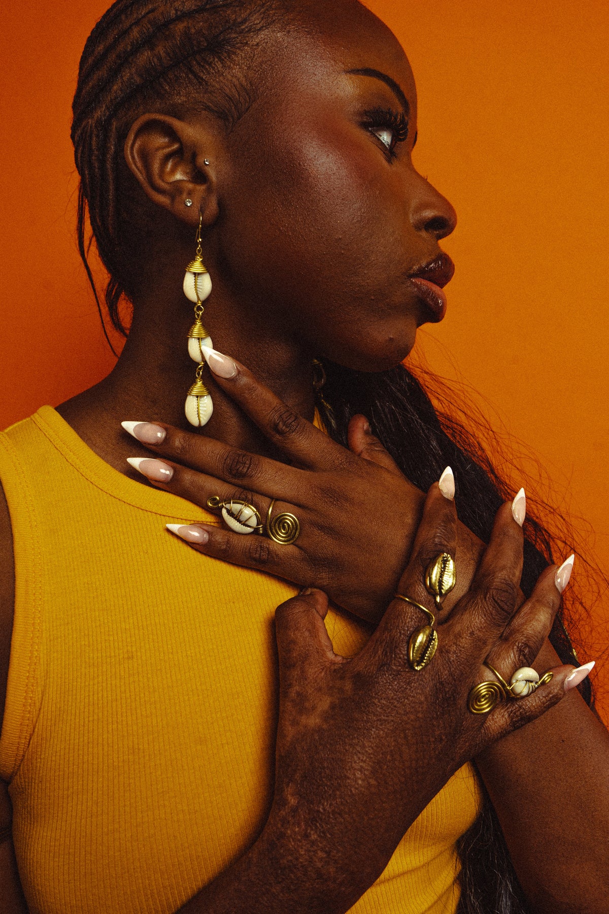 Femme de profil sur fond orange, portant des pendantes à trois cauris sur laiton doré et plusieurs bagues en cauris et spirales de laiton