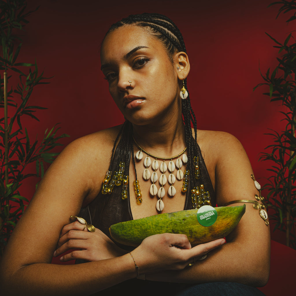 Femme aux tresses portant le collier de cauris à plusieurs rangs, une papaye verte dans les bras, sur fond rouge