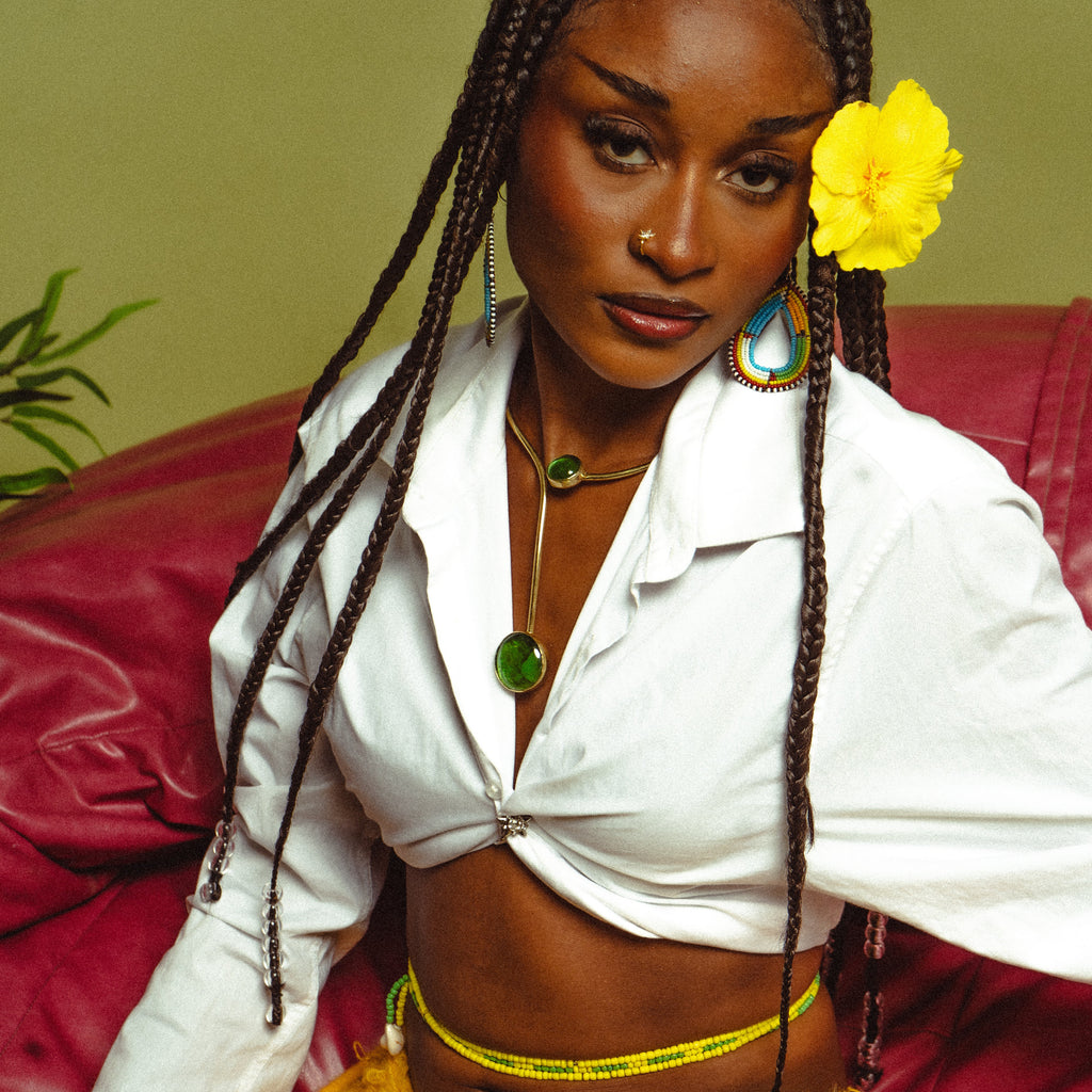 Collier asymétrique à pierres vertes porté avec une chemise blanche, une fleur d'hibiscus jaune dans les tresses