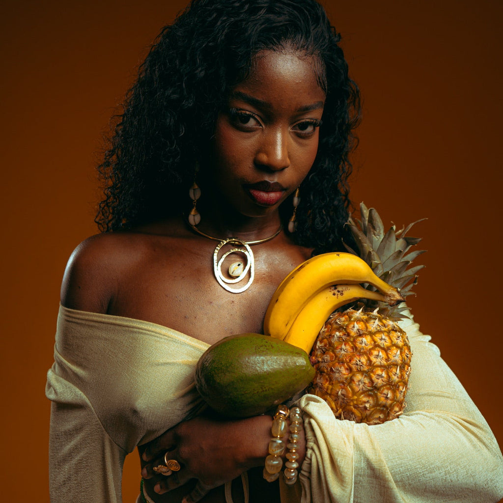 Collier à pendentif spiralé doré porté sur un haut crème, ananas, bananes et avocat dans les bras, sur fond ocre