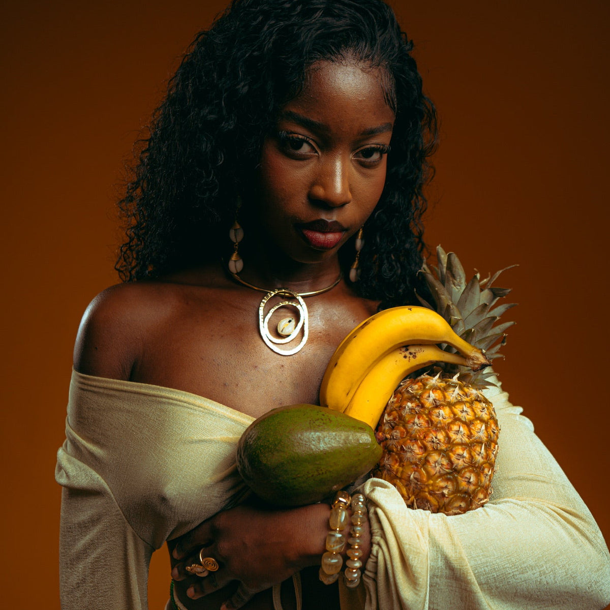 Collier à pendentif spiralé doré porté sur un haut crème, ananas, bananes et avocat dans les bras, sur fond ocre