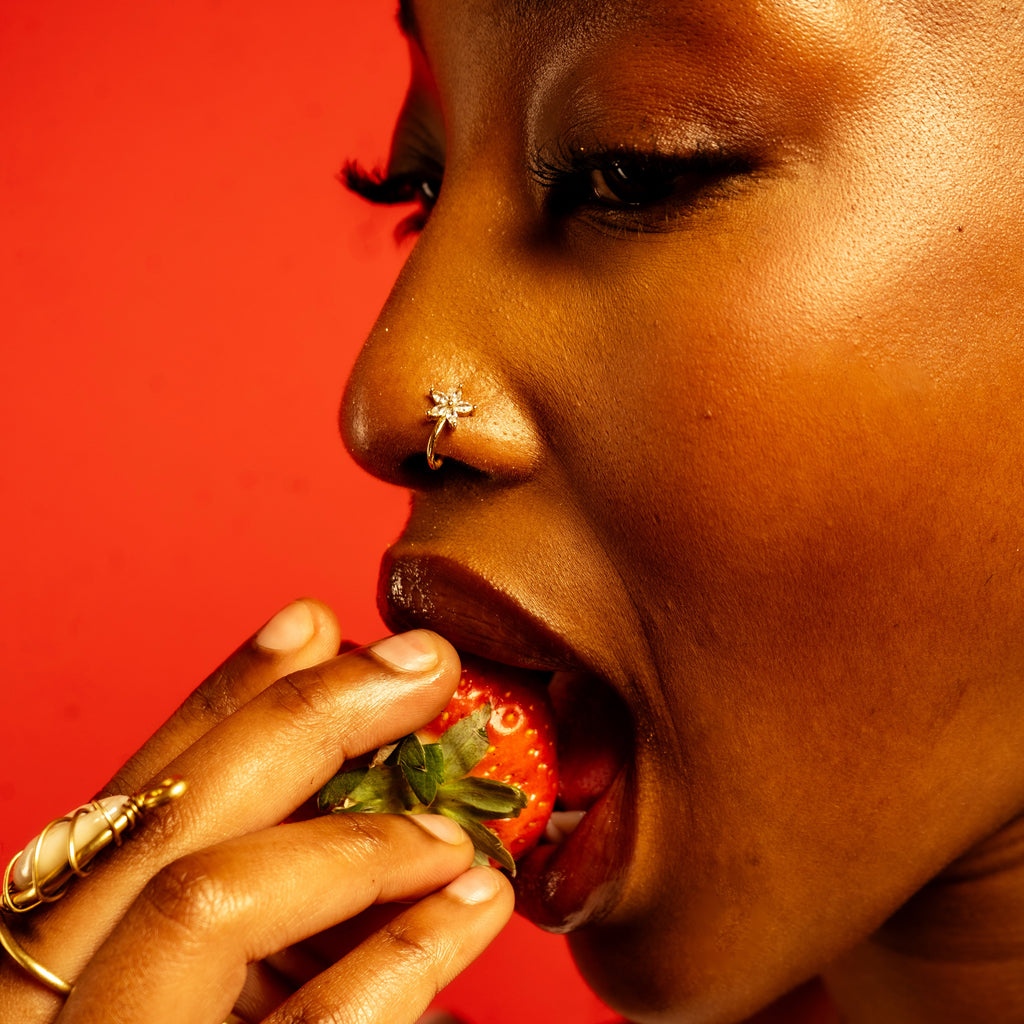 Piercing de nez en fleur dorée porté de profil, une fraise à la bouche, sur fond rouge