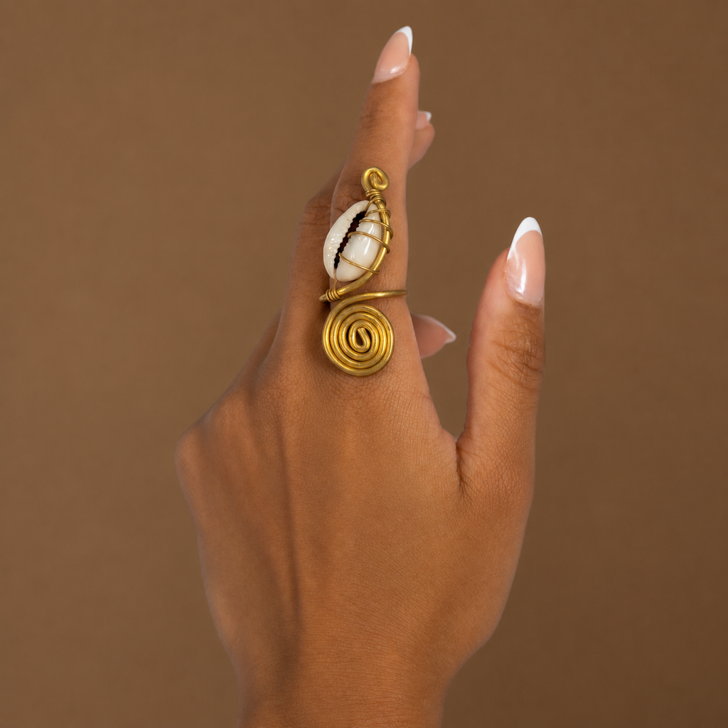 Bague spirale en laiton doré ornée d'un cauri blanc, portée à l'annulaire, sur fond brun