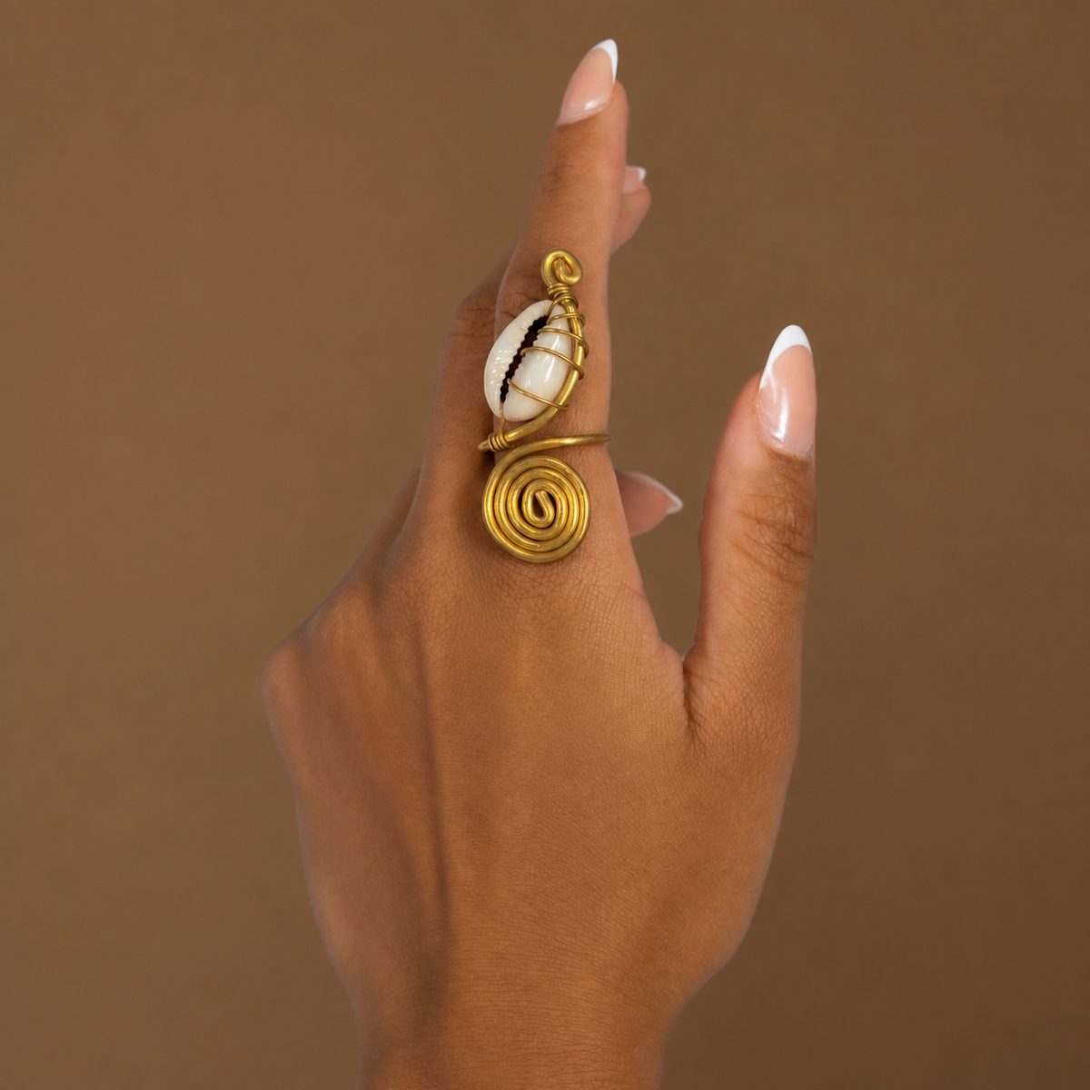 Bague spirale en laiton doré ornée d'un cauri blanc, portée à l'annulaire, sur fond brun