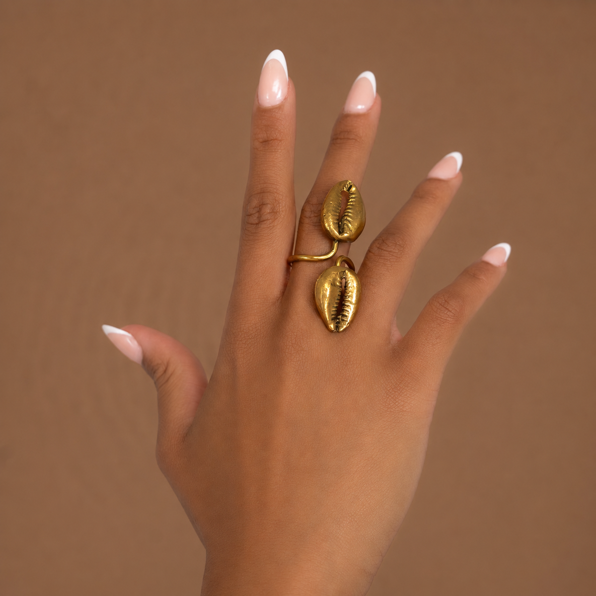 Bague en laiton doré à deux cauris dorés, portée sur deux doigts, sur fond brun
